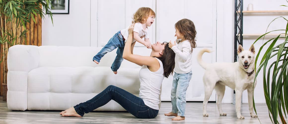 Young mom sitting on floor holding child up in the air while other child and dog stand by.