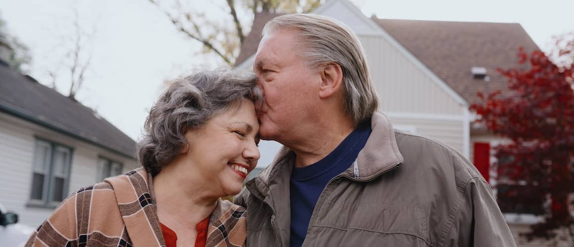 Man kissing forehead of smiling wife in front of home.