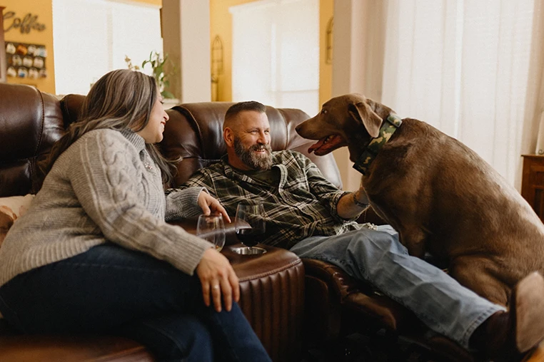 Man and woman relaxing on leather chairs in a living room, smiling at each other, with a brown dog standing nearby.
