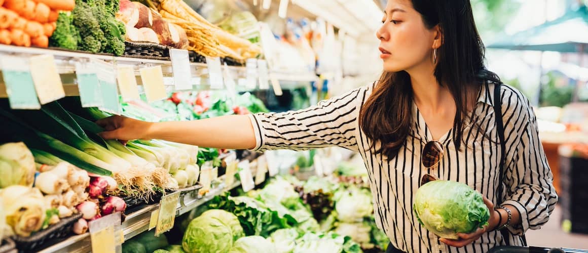 Young woman standing in produce aisle, holding lettuce and reach for more green veggies.