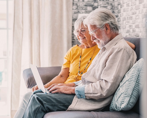 A senior couple sitting side by side on a couch together while using a laptop.