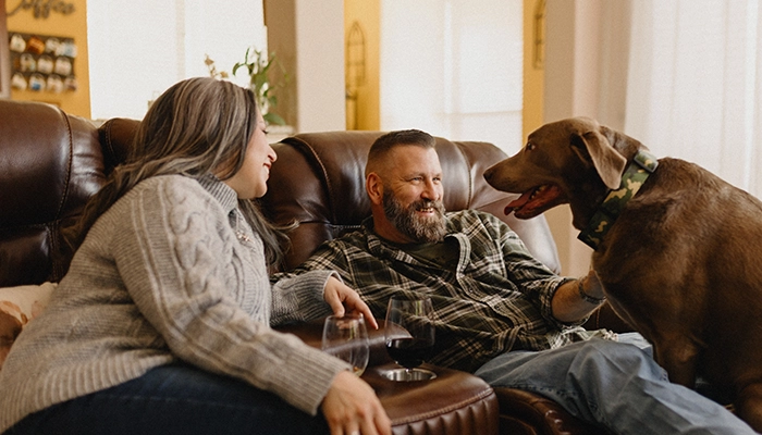 Couple sitting on brown couch while brown large dog climbs up.