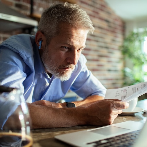 Man in blue shirt hunching over table, holding paperwork and looking at a computer.