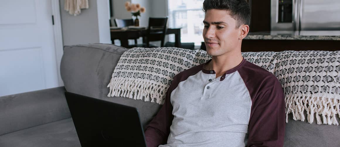 A young man sitting on his home couch looking at something on his laptop.