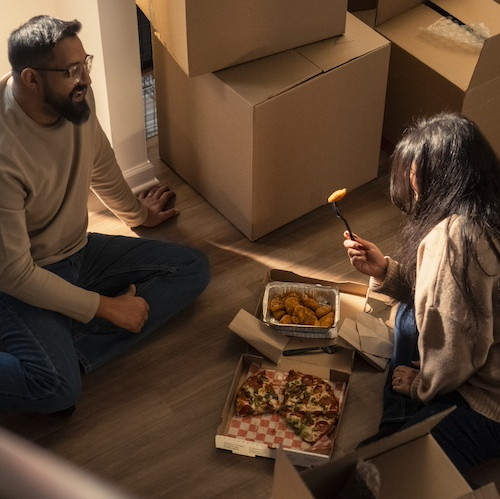 Man and woman sitting on floor of new home surrounding by boxes, eating take out.