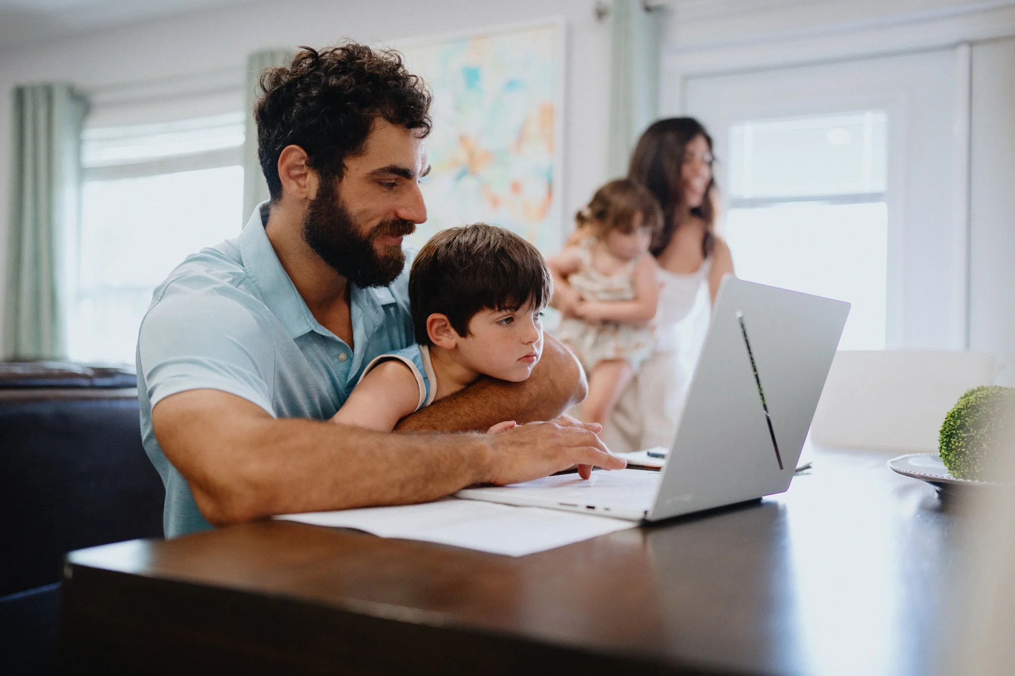 Man working on a laptop at a table while holding a child, with family members nearby in a bright living space