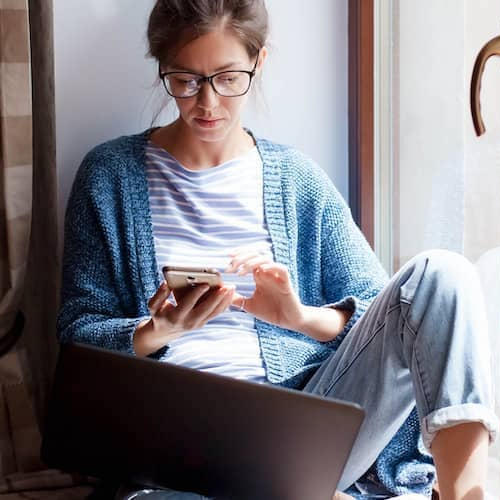 Woman checking credit score via phone on home window seat.