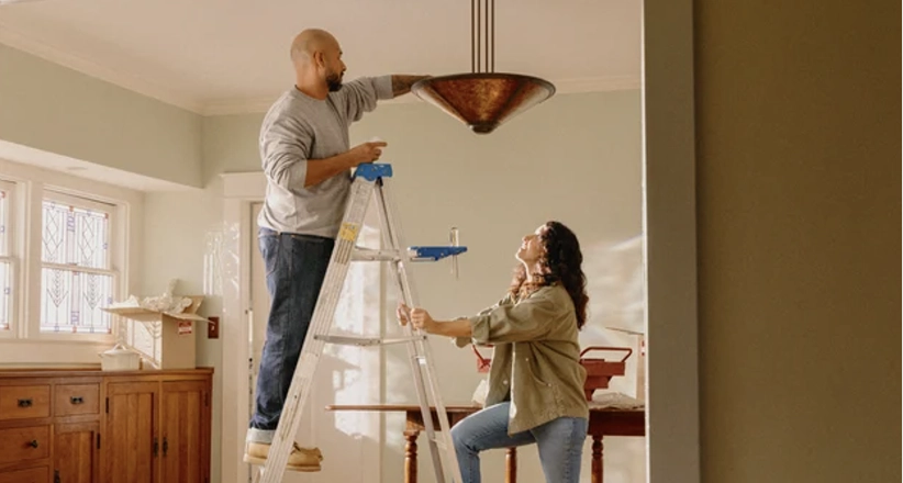 A man stands on a ladder adjusting a ceiling light while a woman holds the ladder steady.