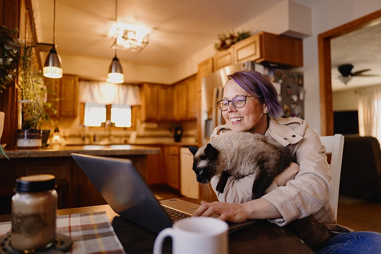 Woman with glasses working on a laptop in her kitchen, smiling with a Siamese cat on her arm.