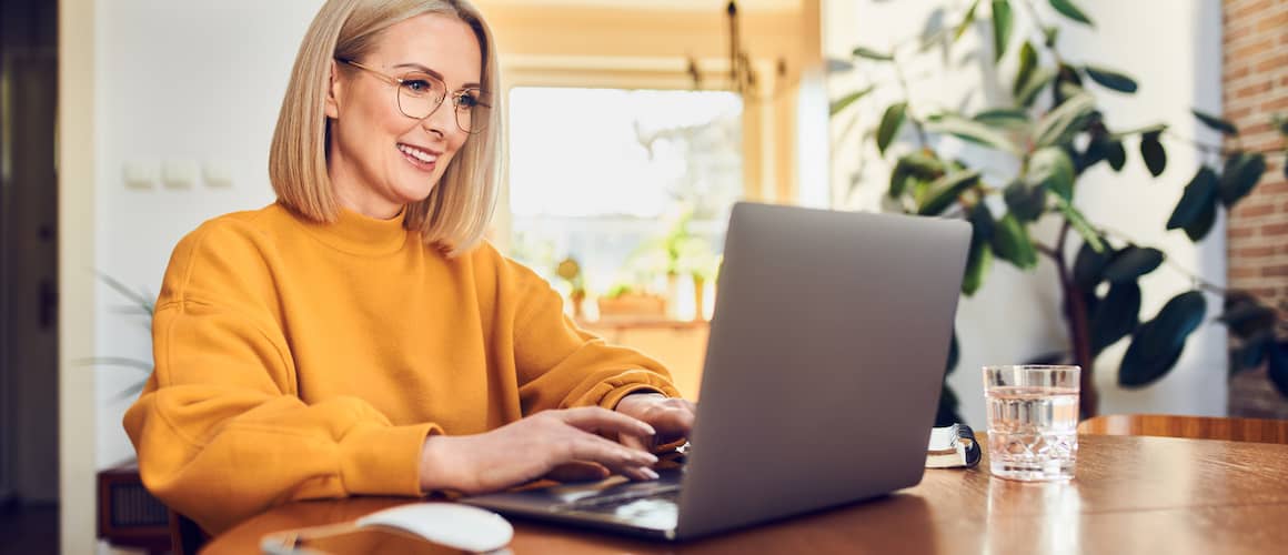 Middle-aged woman wearing glasses and orange sweatshirt, sitting at table working on computer.