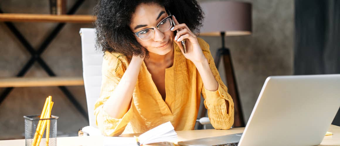 Woman looking pensive on phone with laptop open at desk.