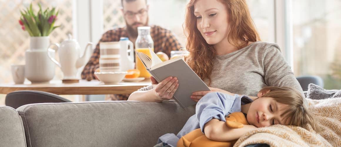 Image of woman reading as sleeping child sits on her lap. Man on phone in background.