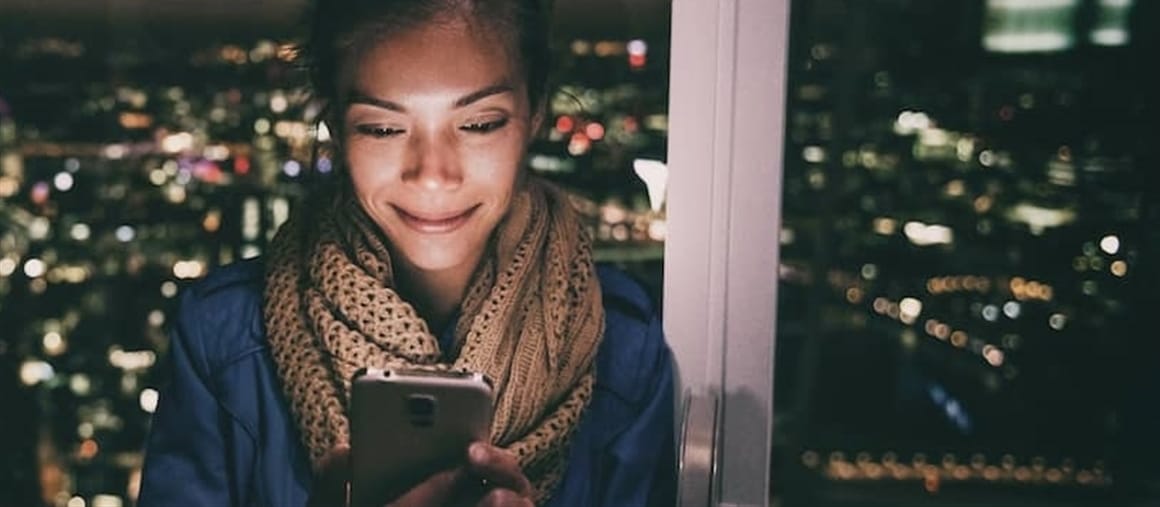 A woman standing in front of a window, engrossed in her phone.