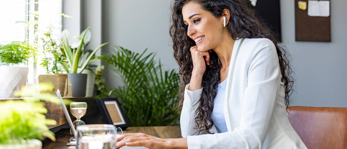 Woman working at desk with earbuds and laptop surrounded by houseplants. 