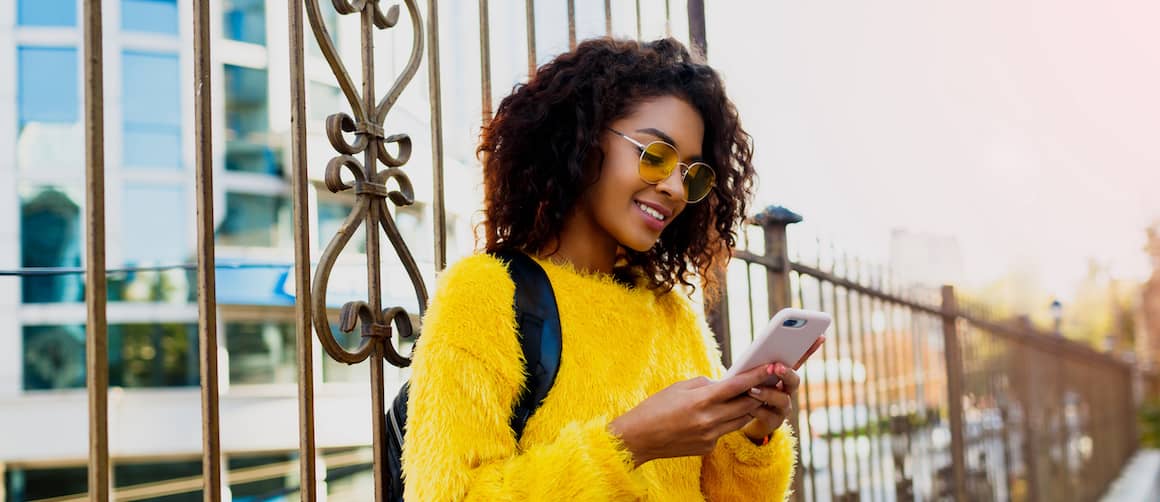 Young woman with glasses looking at phone while leaning against fence.