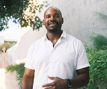 An African American man standing in a park surrounded by trees and talking.