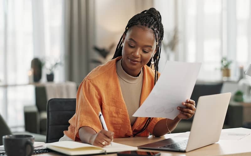 Woman in orange top writing in journal with laptop in front of her.