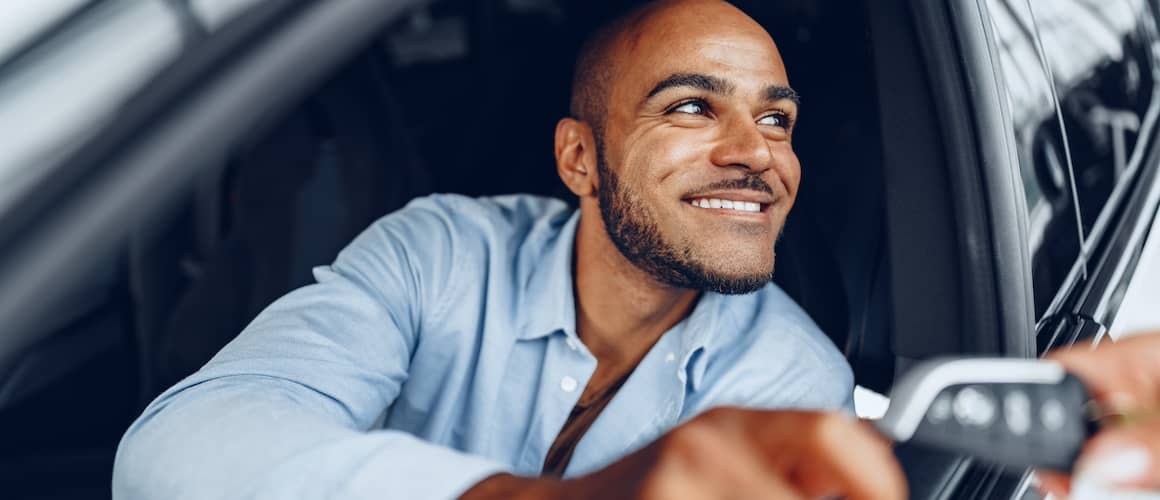 Young smiling man in drivers seat of new car being handed keys by salesperson.