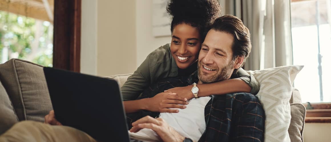 Couple snuggling on couch and reviewing information online on open laptop.