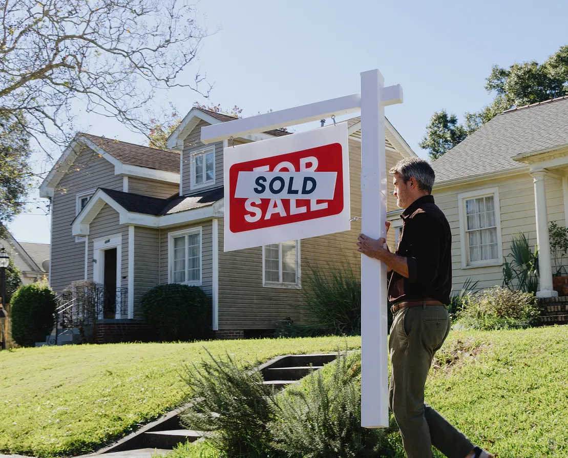Person standing beside a sold sign in front of a home