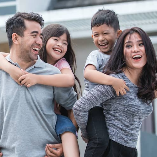A couple with two kids giving piggyback rides in front of their house, symbolizing family happiness and homeownership.