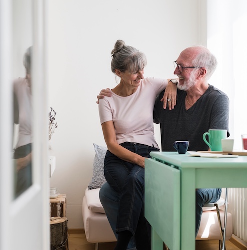 Older couple smiling and snuggling at small jade green kitchen table while having coffee.