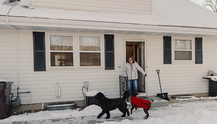 Large black dogs play in snow of backyard while woman holds door open.