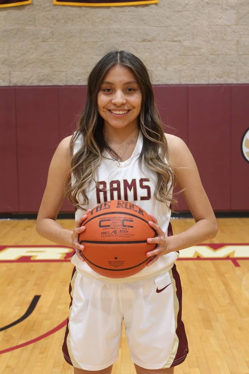 Girl posing with basketball, in while basketball uniform, smiling.