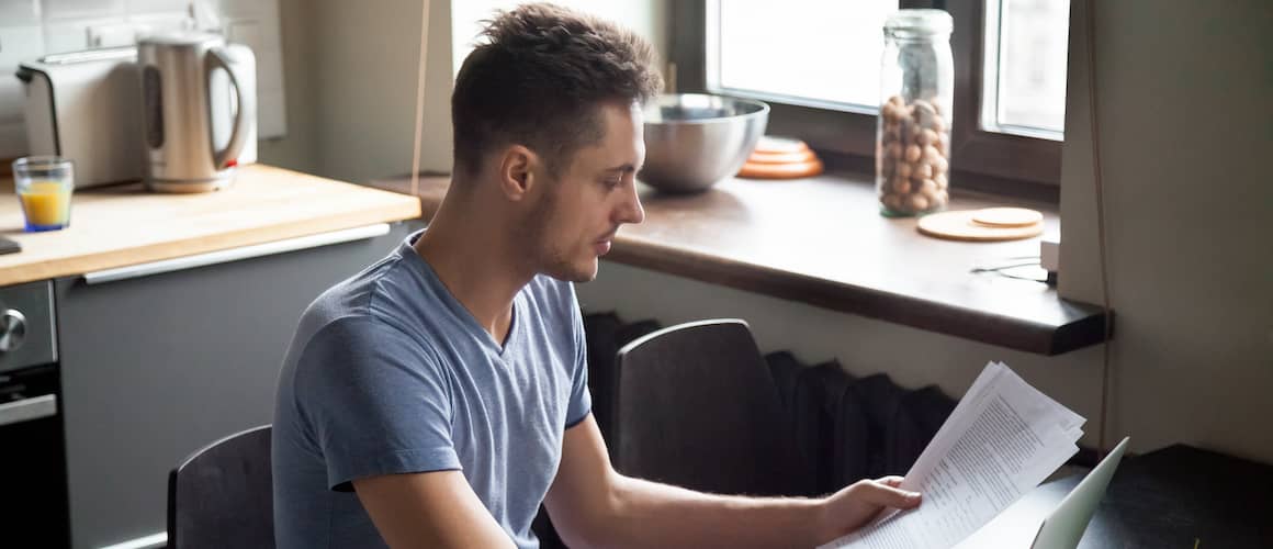 Young man in kitchen at home using a laptop and holding papers.