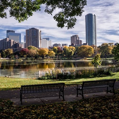 City view of Minneapolis, Minnesota from a park with two benches and two geese along the edge of the water.