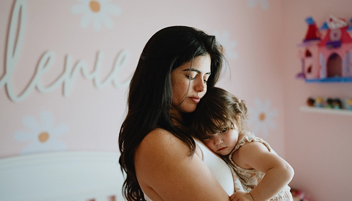 Woman holding toddler in pink baby room with whit crib in background.