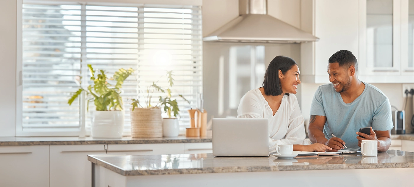 A Black couple enjoying morning coffee at their kitchen counter smile at each other while working on their home buying plans.