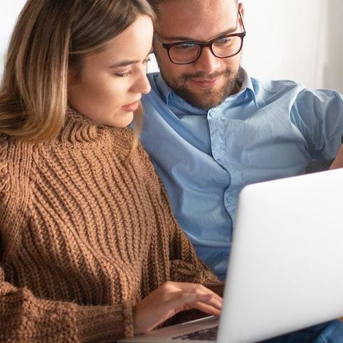 Smiling young couple using silver laptop while green houseplants sit on the wooden table behind them.
