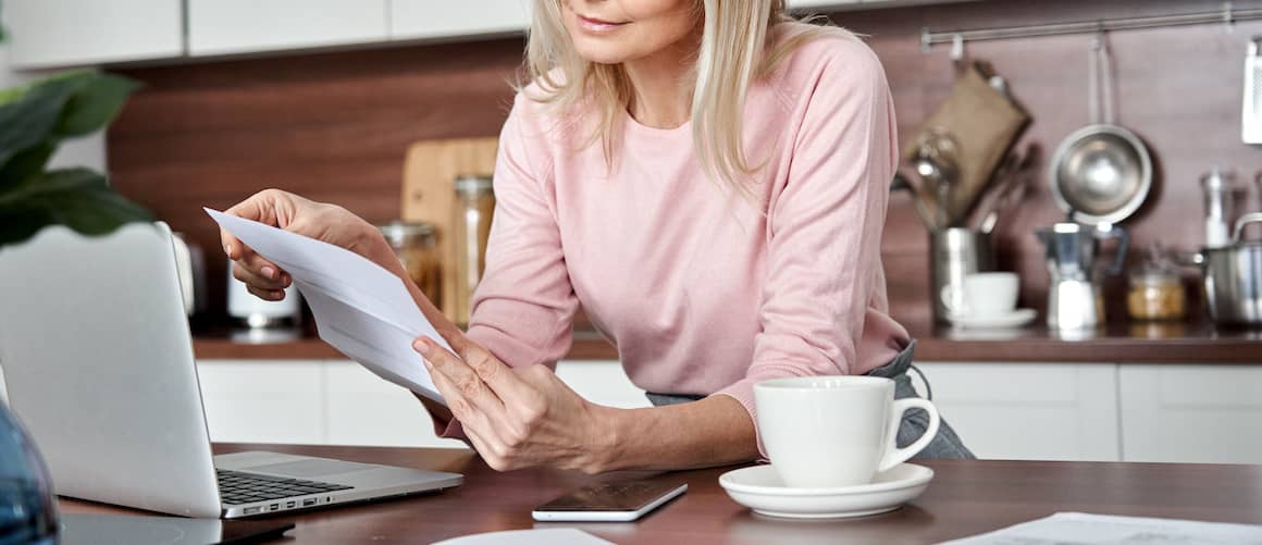 Woman reviewing invoices at kitchen island with open laptop, coffee cup nearby.