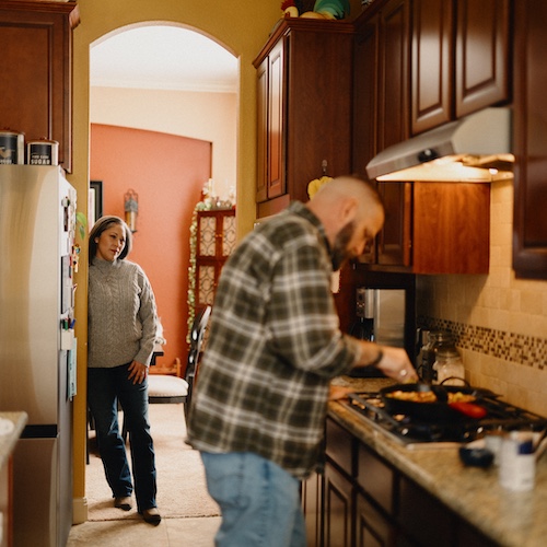 Woman watching man cook in kitchen with warm-toned cupboards.