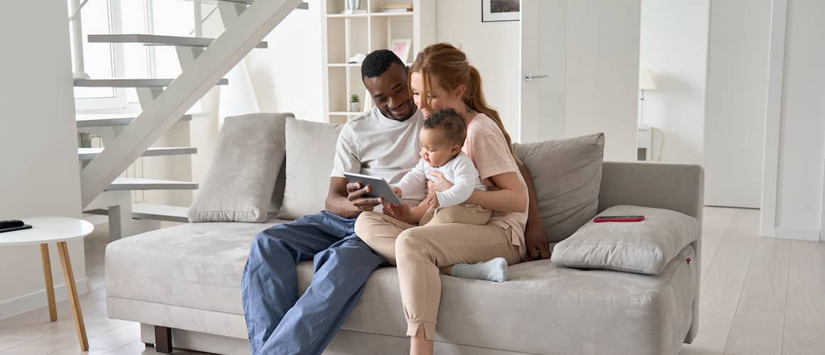 Parents and their young child sit on a couch in a living room and look at their phone or tablet.