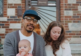 A Black man wearing a baseball cap and glasses holds his six-month old on his lap while sitting on his front porch with his partner, a white woman with long hair gazing downward