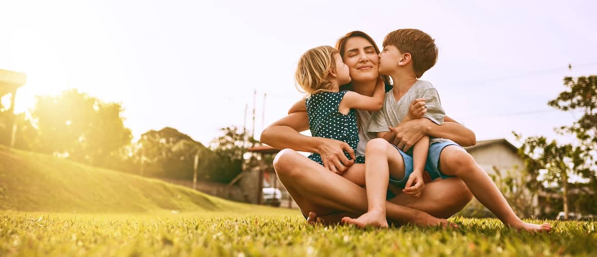 Mother sitting on grass with two children on her lap, kissing her cheeks.