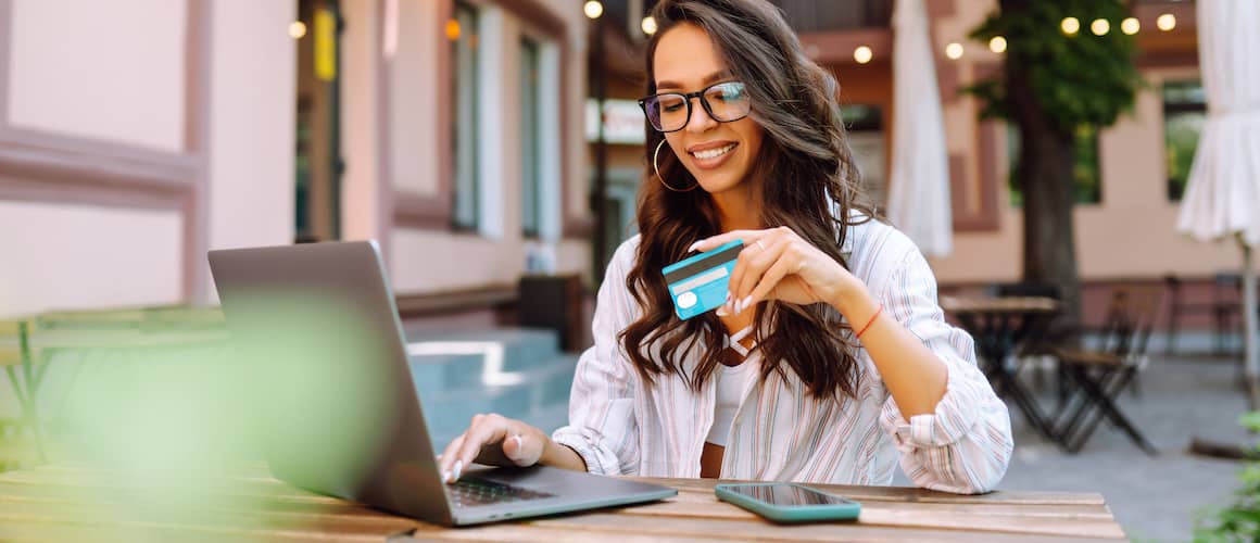 Young woman sitting outside at a wooden table and bench using her credit card through a laptop.