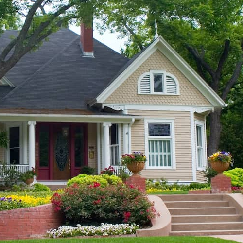 Tan house with a double-layered front lawn, brick steps, and a wrap-around porch.