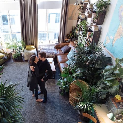 Couple hugging in condo living room with large windows surrounded by houseplants.