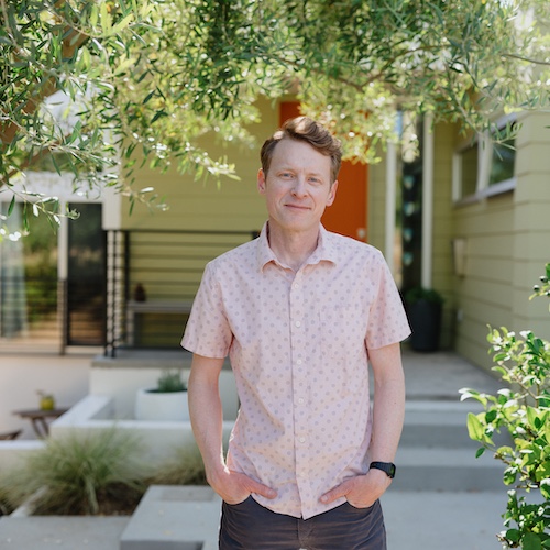 Man in salmon colored button up smiing in front of home under trees.