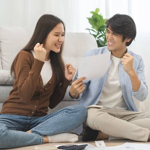 Young couple sitting in front of a couch and excitedly looking over paperwork.