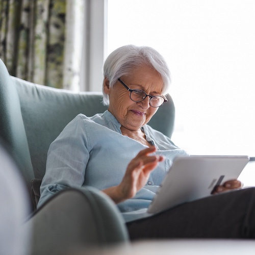 Senior woman sitting in a light blue wingback armchair while using a tablet.