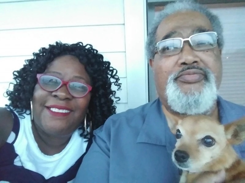 Woman and man on their front porch smiling, holding a small brown dog.