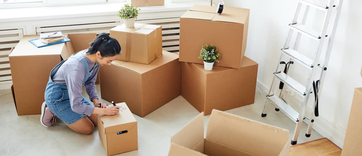 RHB Assets From IGX: Young Asian woman labeling cardboard boxes in a bright, white room during a move.