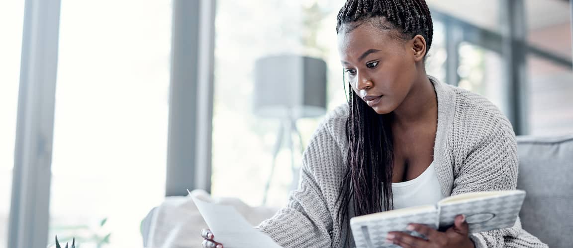 Young woman sitting on couch, holding notebook and reviewing budget document.