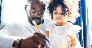 Hombre negro escribiendo algo mientras su niño pequeño está a su lado.