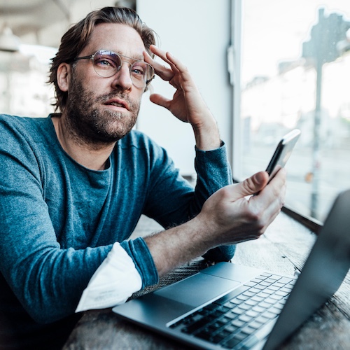 Man holding hand to head, looking into the distance and holding his phone with his laptop open nearby.