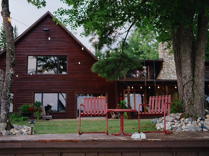 Dark red house with string lights, stone chimney, and two red chairs with a table in the yard.